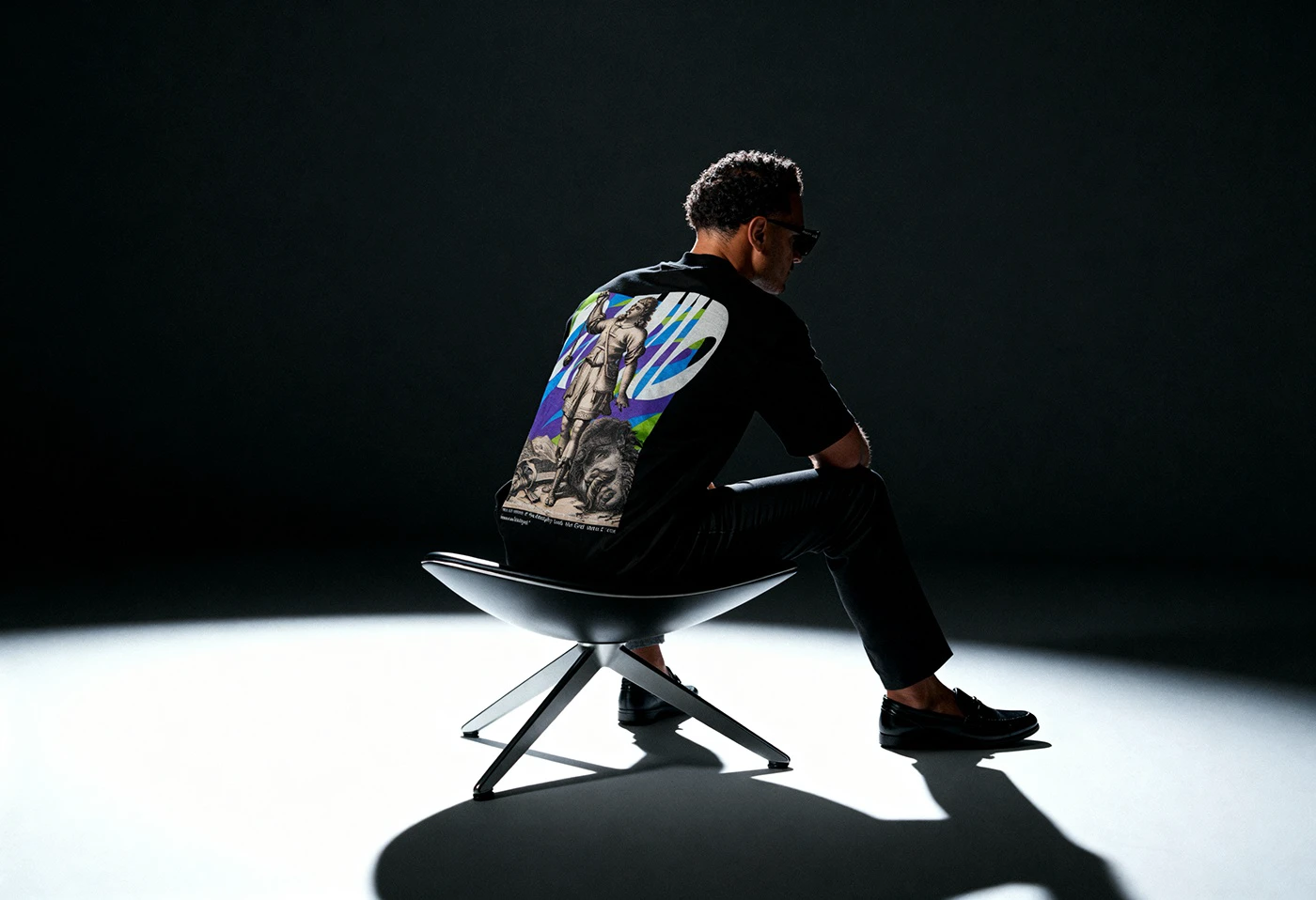 Model sitting in a dark studio, showing the back print of the black Timeless tee